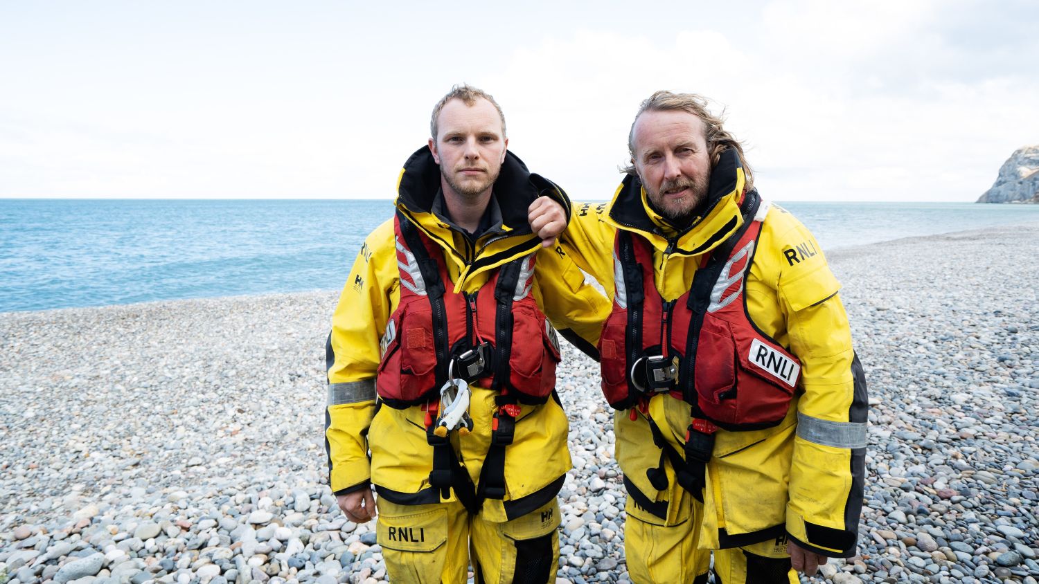 Graham and Luke Heritage stand on the beach in their RNLI kit looking serious.