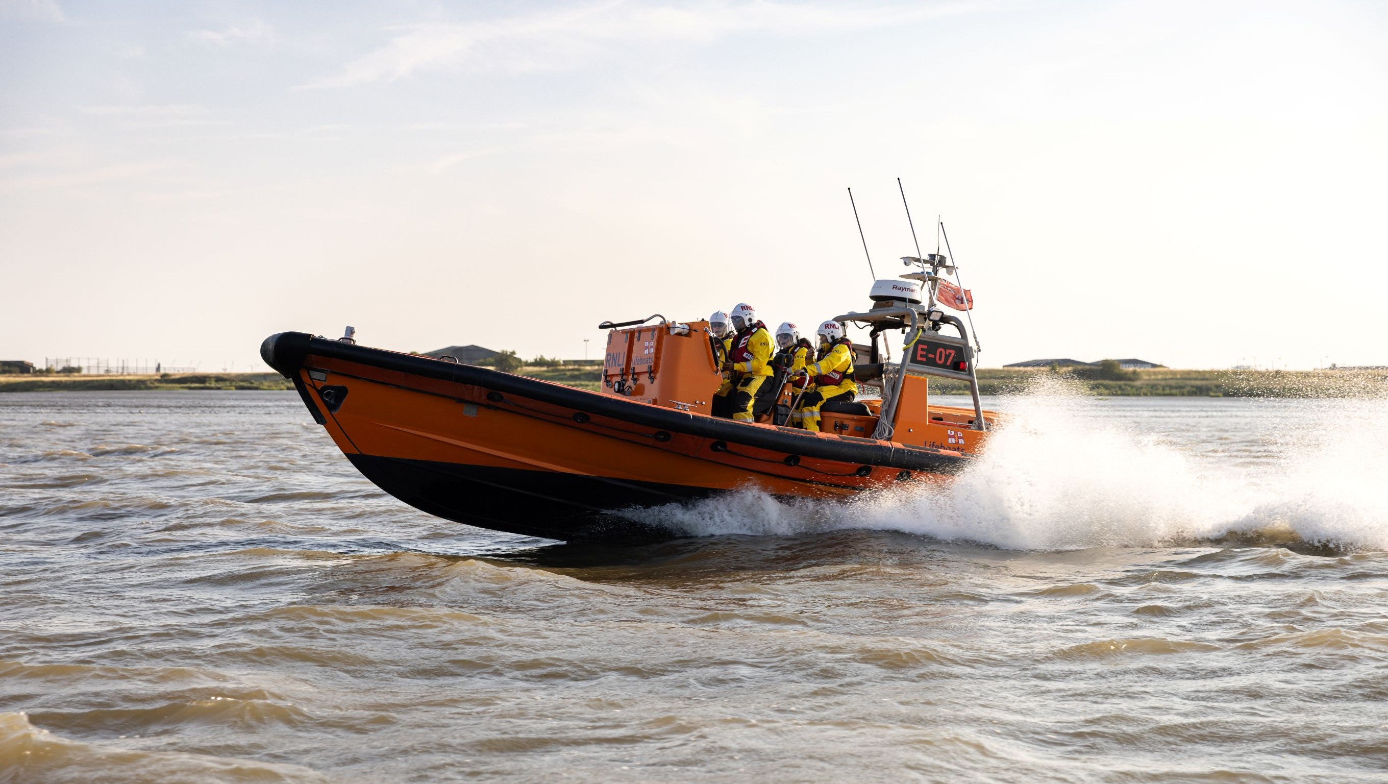A side view of an E class lifeboat and its wake as it powers through the water on the River Thames with four crew members on board.
