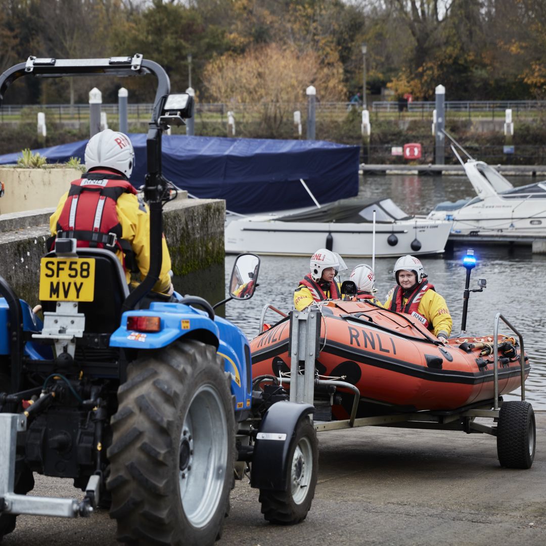 Teddington RNLI's D class lifeboat being launched down the slipway by tractor and trailer. Three crew members are in the lifeboat and one is driving the tractor. 