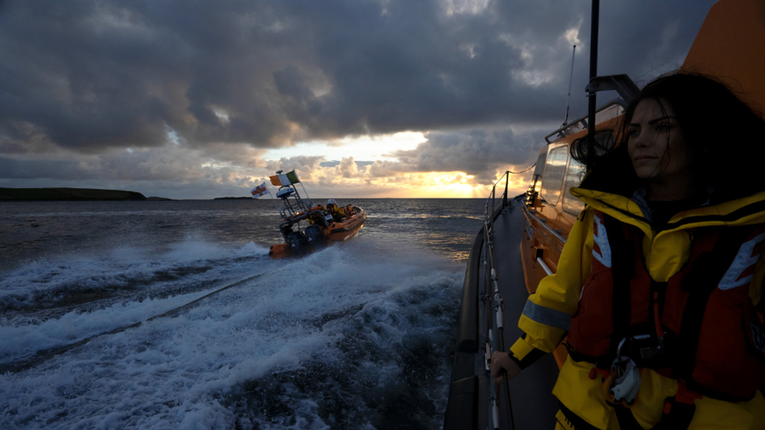 A female crew member stands on the deck of Clifden’s Launch a Memory Shannon class lifeboat as the station’s B class lifeboat cruises past. It’s sunset and light breaks through between darkening skies and grey seas. 