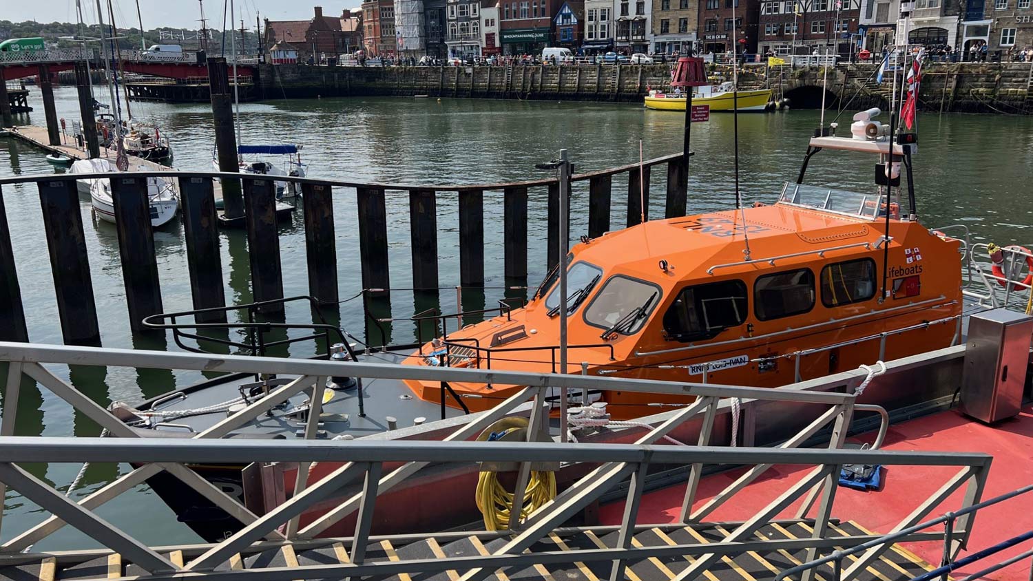 Whitby’s Shannon class Launch a Memory lifeboat sits moored in the harbour next to a gangway, with the harbour, sea wall, and busy quay