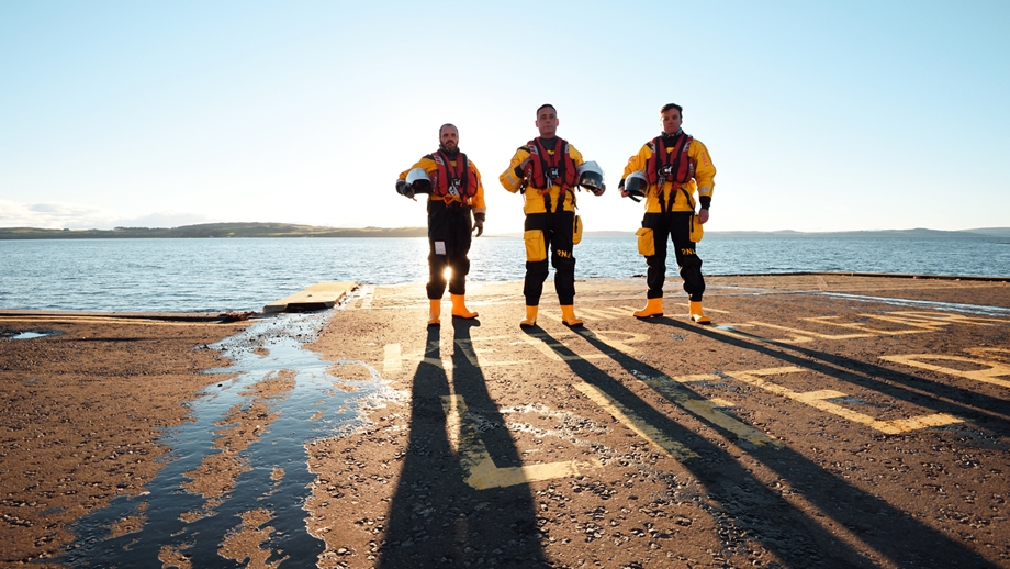 3 male RNLI crew members stand next to a body of water