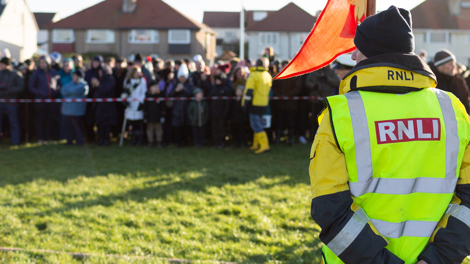 A man in a high-vis RNLI jacket has his back to the camera, as though an event is about to begin. Flags wave. A person in lifeboat crew kit is asking the watching crowd for donations to a collection bucket.