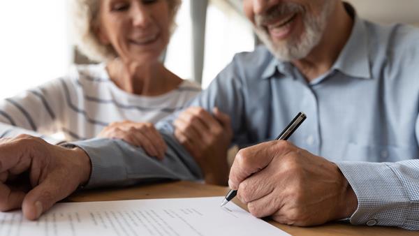An elderly couple sitting together, smiling warmly. The man, wearing a light blue checkered shirt, is signing a document with a black pen, while the woman, dressed in a white and grey striped top, rests her hand on his arm in a supportive gesture. The focus is on the man's hand and the document, with both appearing content and engaged in the process.