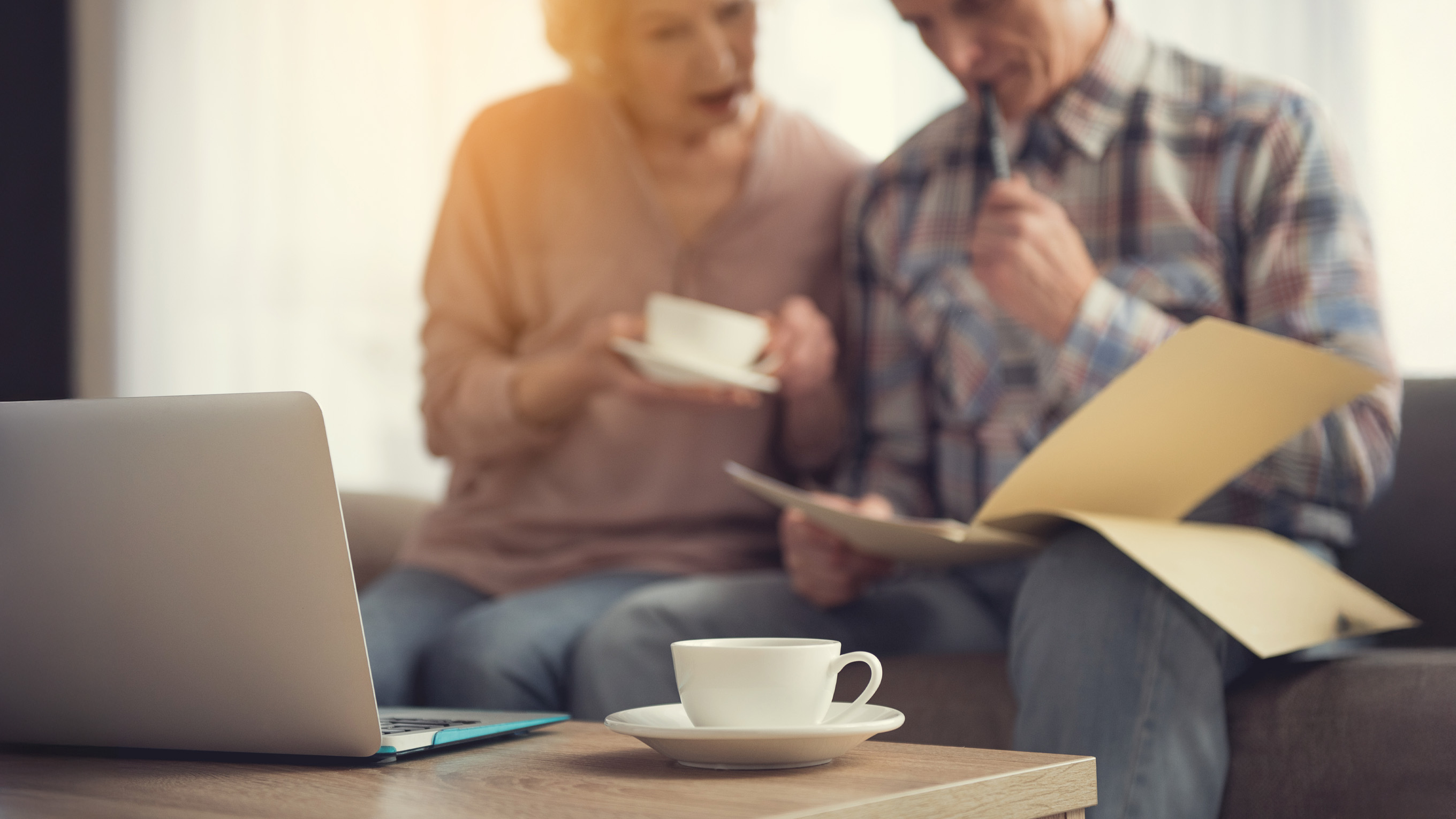 A lady in a pink jumper and a man in a check shirt sit on a sofa reviewing a file of documents with a laptop open on a coffee table in front of them. 