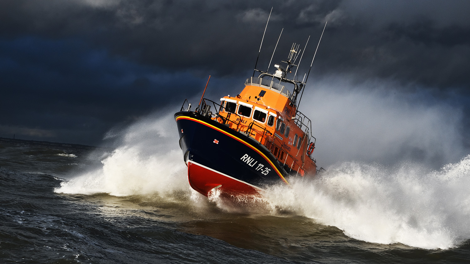 Yarmouth Severn class lifeboat is powering through the sea on a grey, stormy day with lots of white spray around it.