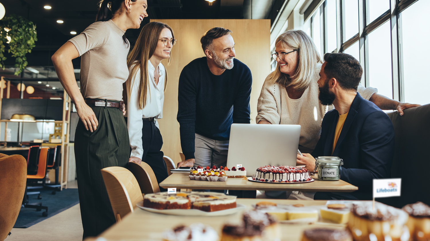 Five smiling people are in an office, standing behind tables full of cakes that are being sold to raise money for charity