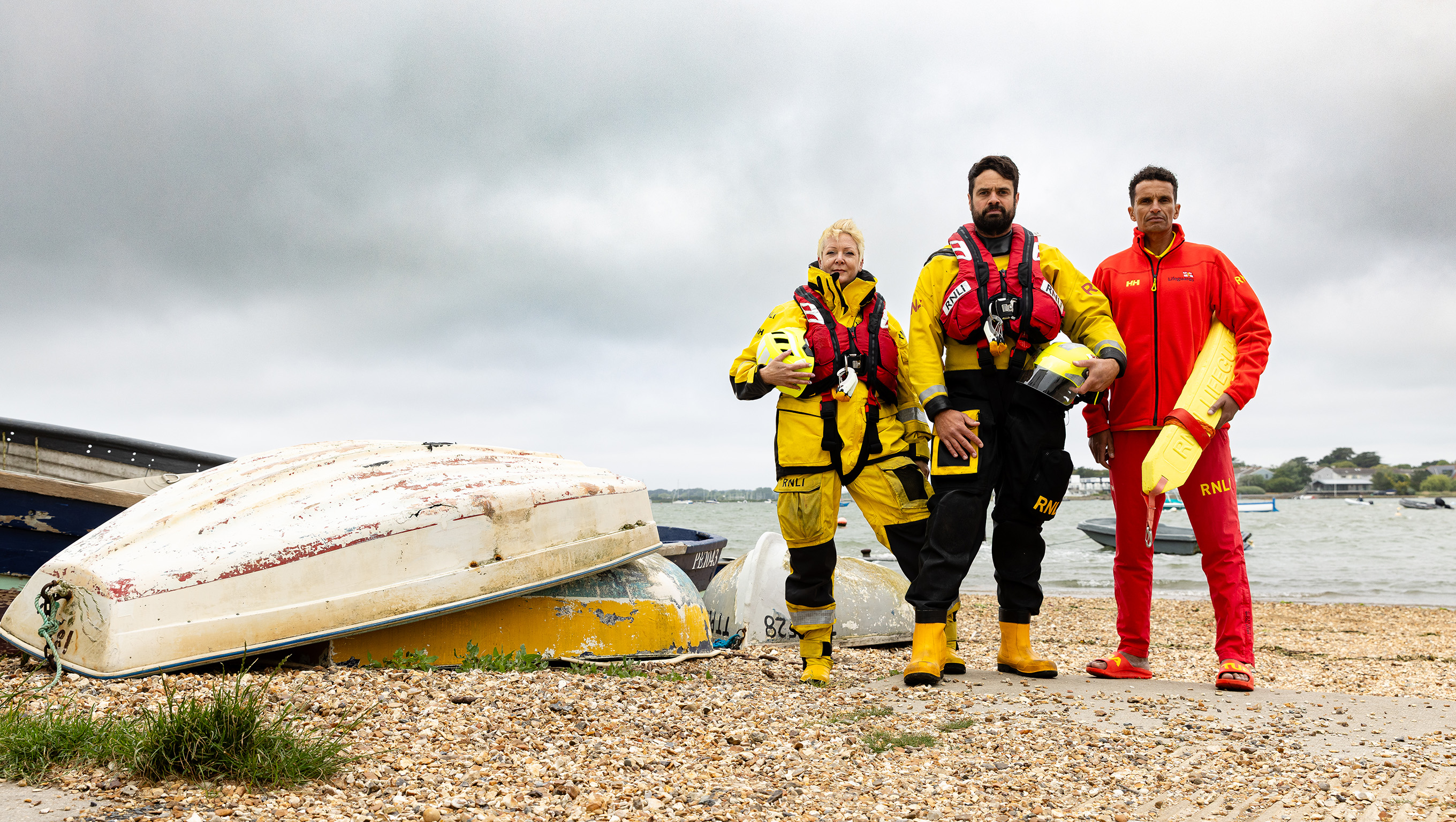 Two lifeboat crew members and an RNLI lifeguard are stood next to small upturned boats on a beach on a cloudy day. 