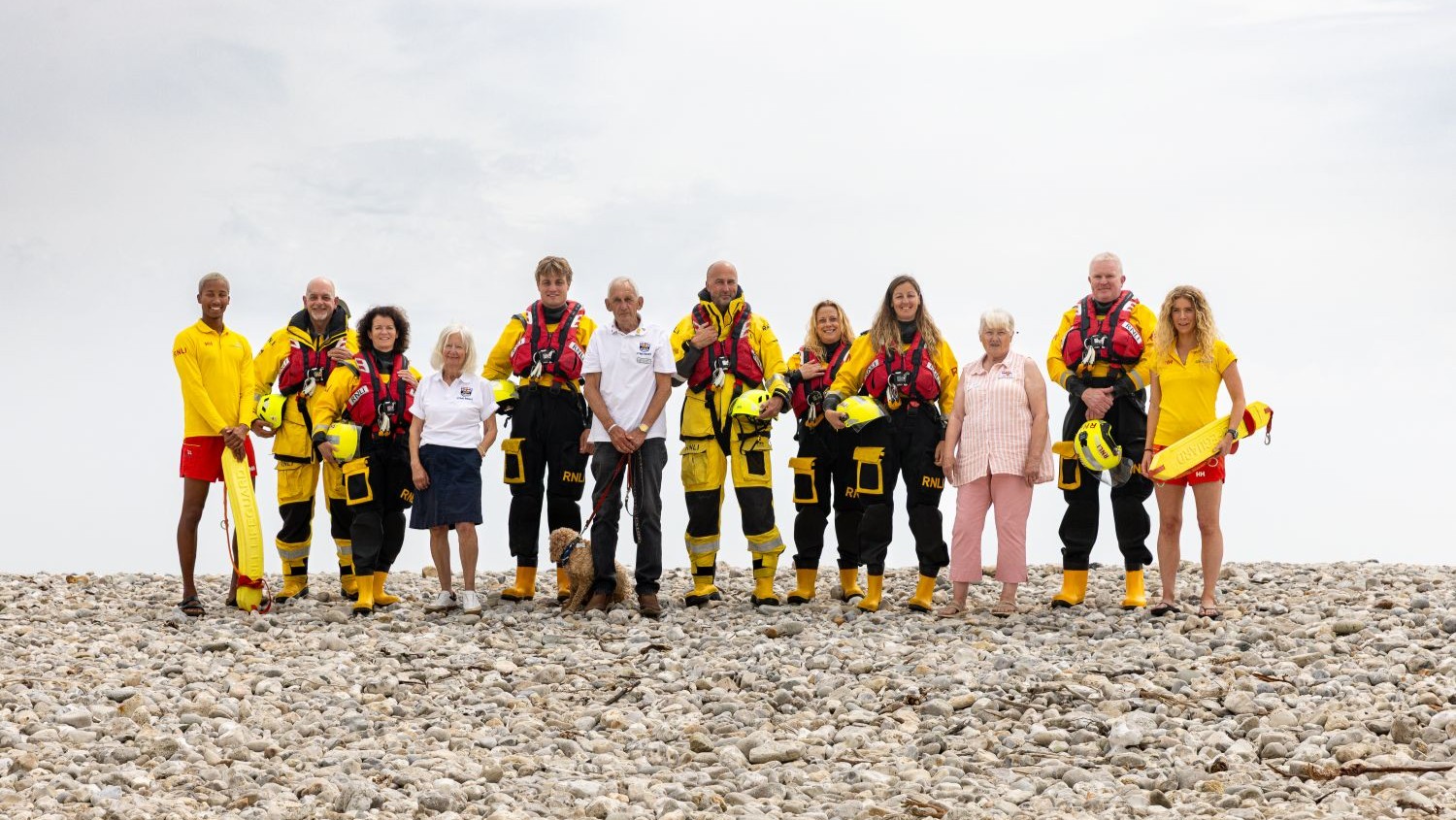 A group of 12 RNLI crew members, lifeguards and shop volunteers stand in a line on a pebble beach, against an overcast sky.