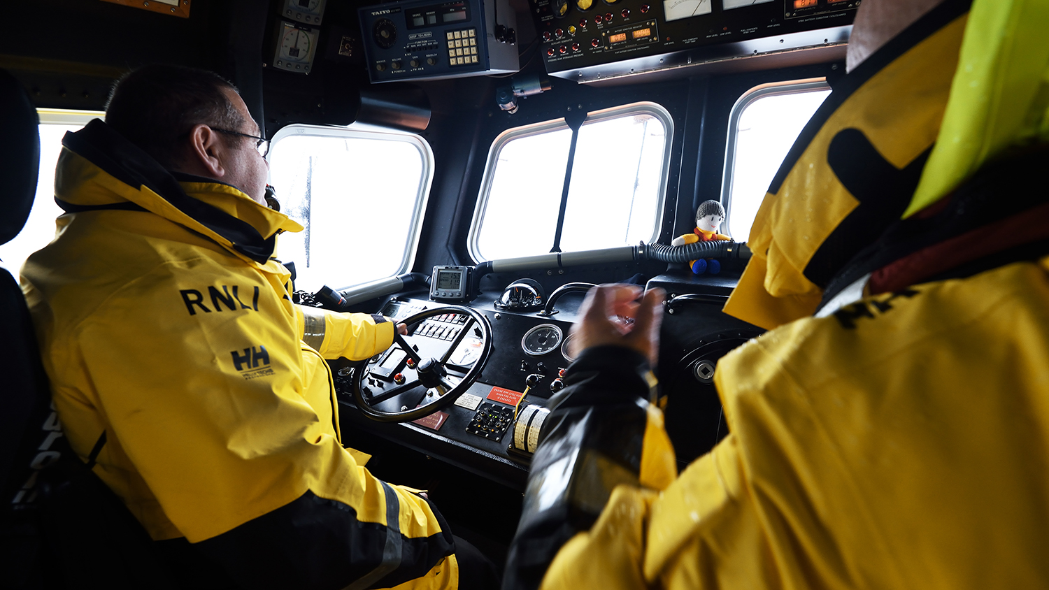 Two crew members are seated inside an all-weather lifeboat, wearing waterproofs. One has his hand on the wheel, steering the boat at sea. There are many controls, buttons and gauges, and windows to the front and to the side of his seat.