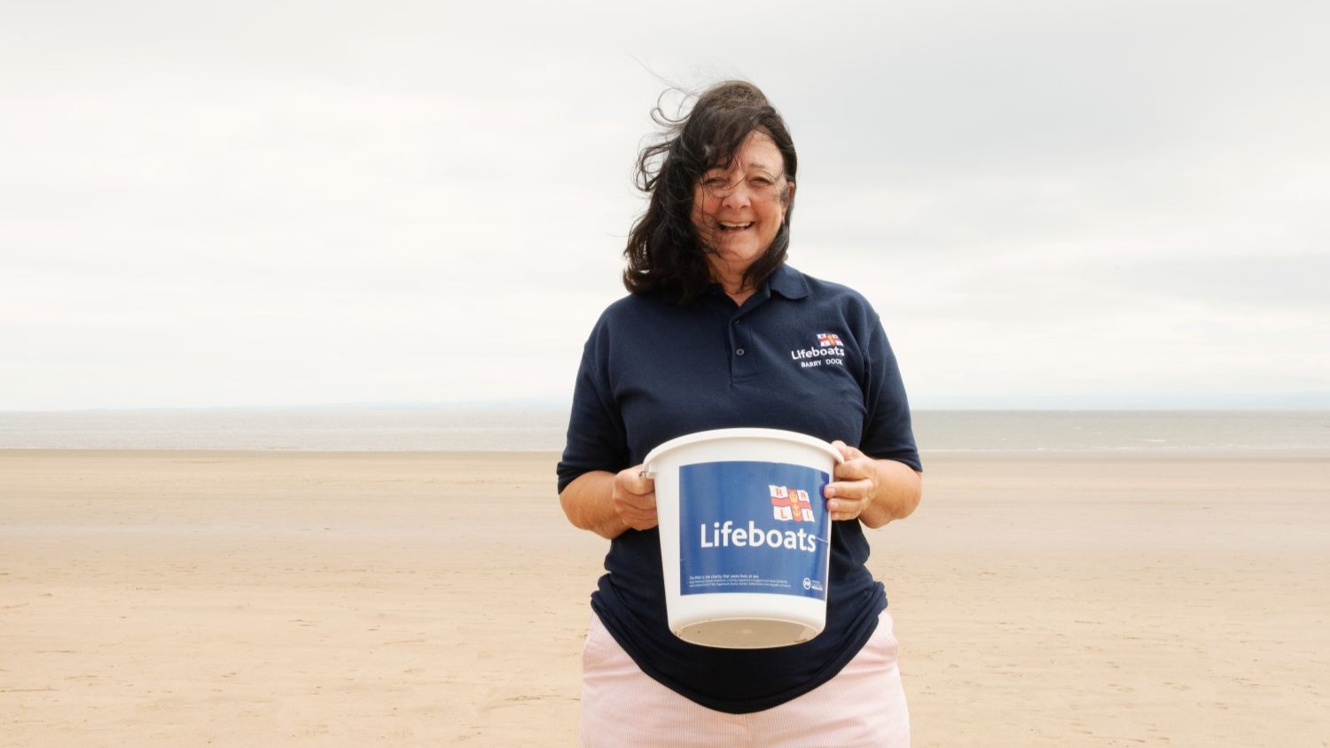 An RNLI volunteer standing on a beach holding a collecting bucket.