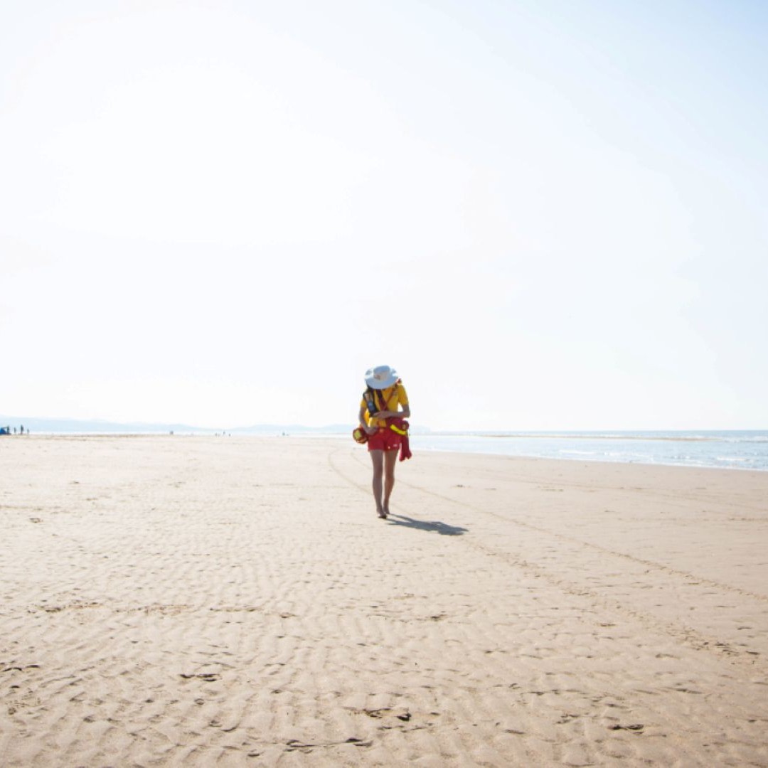 A lifeguard walking alone along a wide, empty sandy beach on a bright, clear day. The sea is visible to the right, with gentle waves along the shoreline, and the sky is pale and cloudless, creating a calm, open atmosphere.