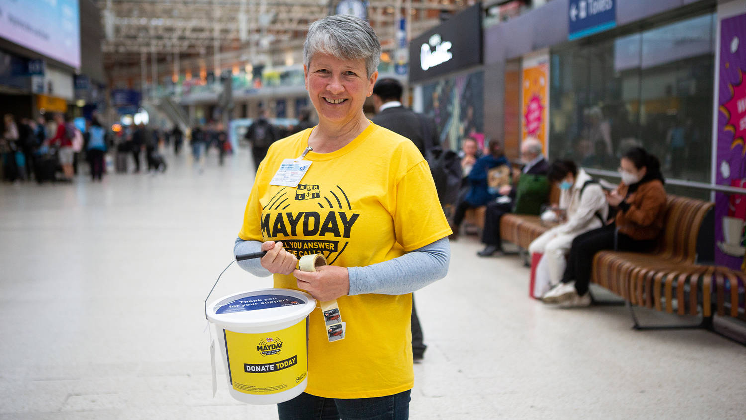 An RNLI fundraising volunteer is stood in a busy train station holding a branded bucket and smiling.