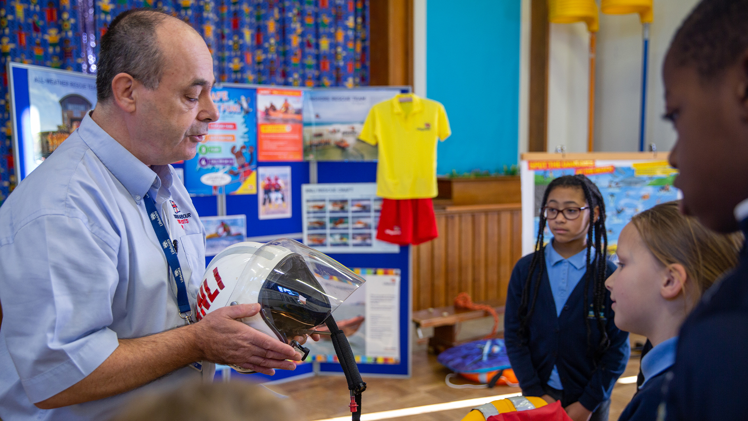 An RNLI water safety education volunteer is holding a lifeboat crew helmet and talking to a group of children in a school.