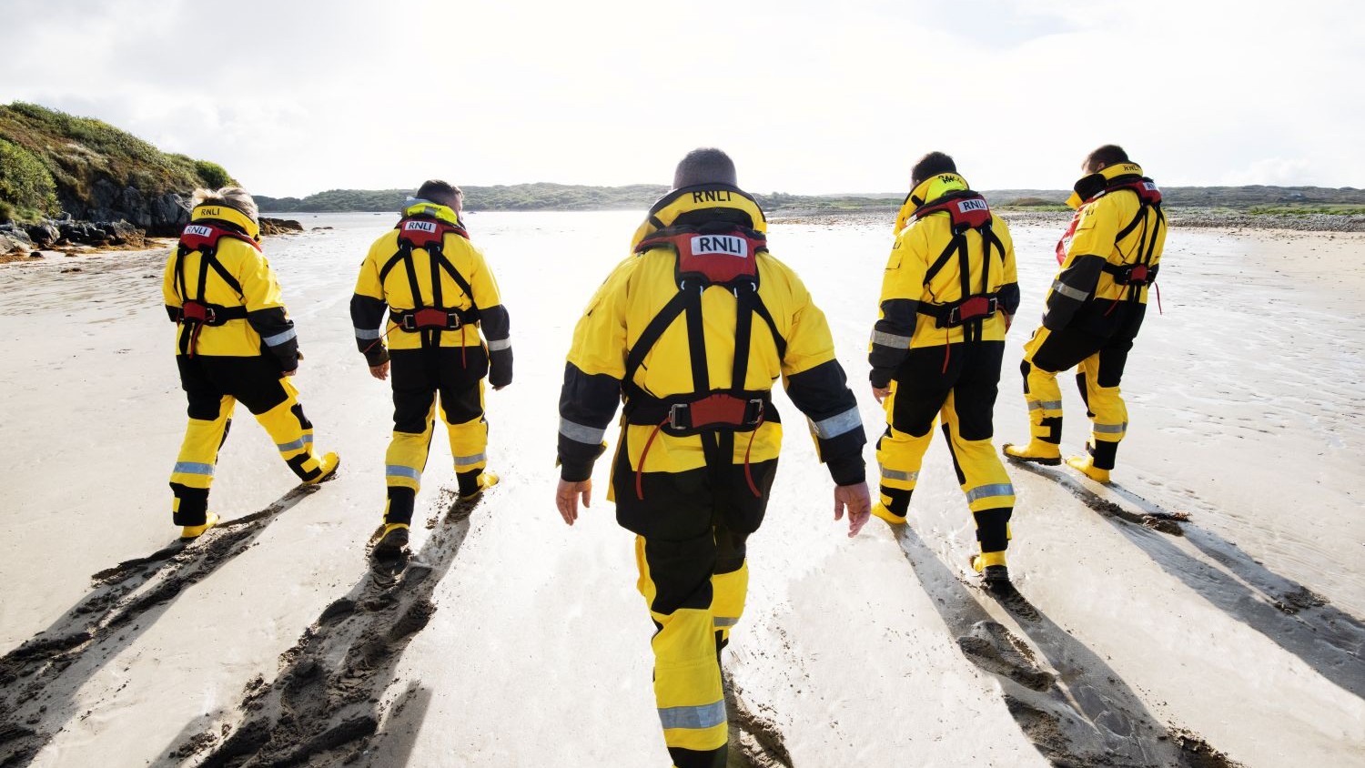 A group of lifeboat crew walk along a beach with their backs to the camera