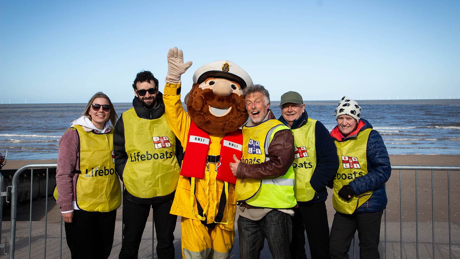 Five people in yellow RNLI Lifeboats vests gather round another figure dressed as the Stormy Stan mascot.