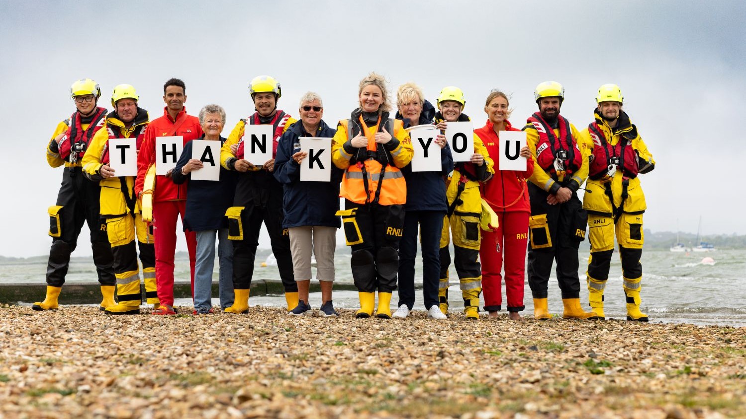 A group of RNLI volunteers standing on a beach. They are holding up cards spelling the word ‘thank you’. 