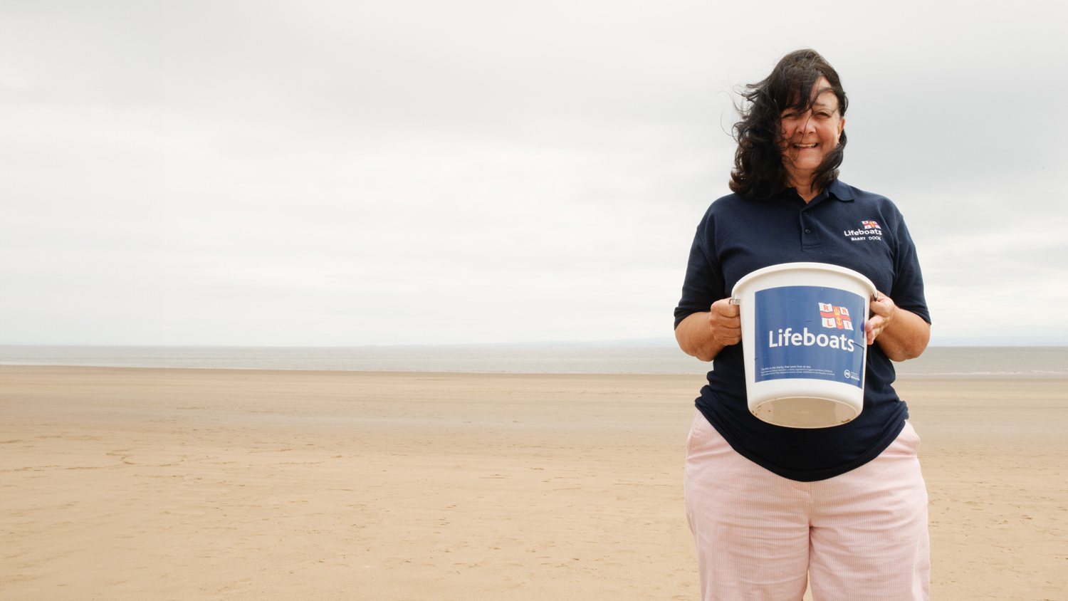 Barry Island Shop Volunteer Avril Kitchen stood on the beach holding a collection bucket.