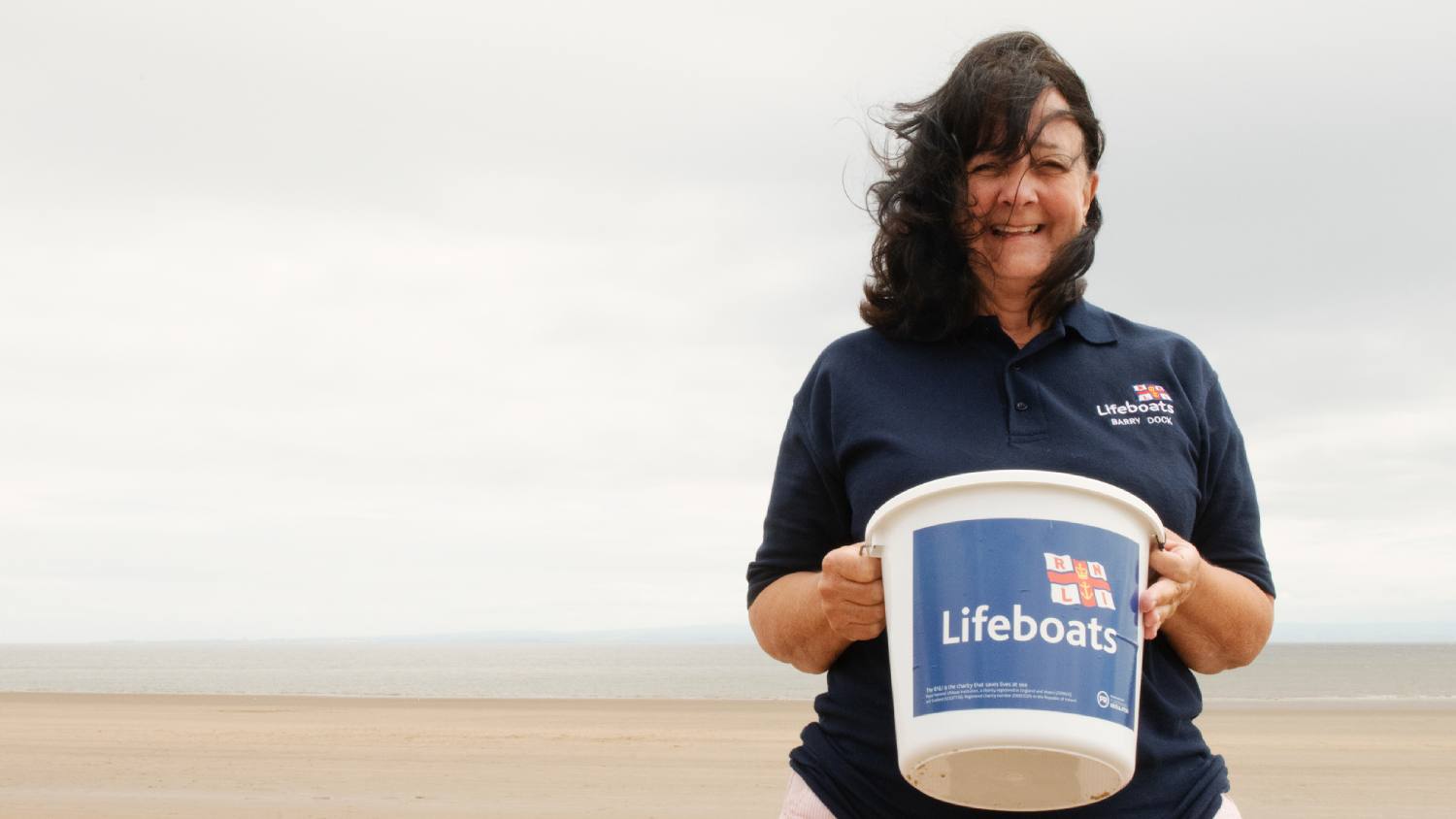Barry Island Shop Volunteer Avril Kitchen stood on the beach holding a collection bucket.