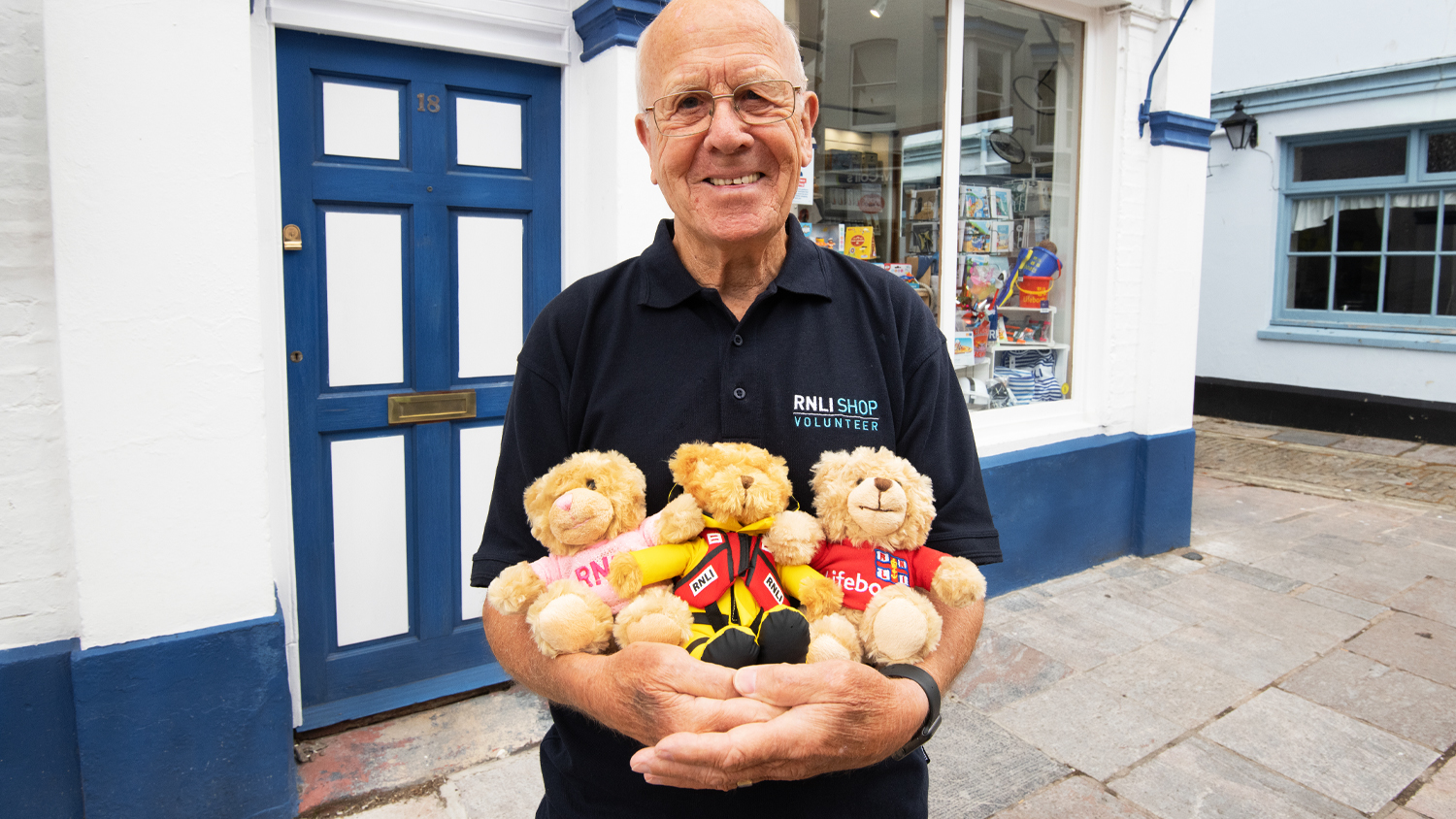A smiling RNLI shop volunteer standing outside an RNLI shop holding three teddy bears.