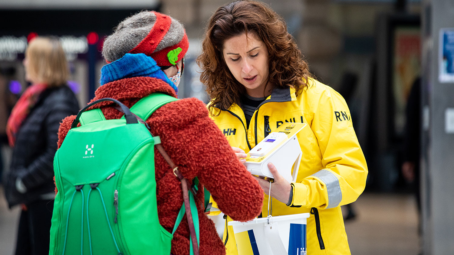 A supporter making a donation with RNLI Fundraising Volunteer and Tower Crew Member Suzanne Goldberg on London Lifeboat Day at Waterloo Train Station. Suzanne is wearing yellow crew kit and is carrying a collection bucket and a contactless payment unit.