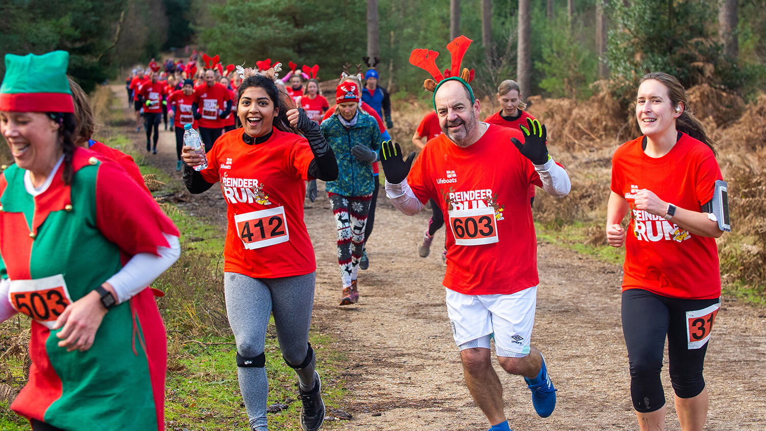 Runners taking part in an RNLI Reindeer Run in Moors Valley Country Park in Dorset. Most runners are wearing red Reindeer Run T-shirts and antlers. Two are waving at the camera.
