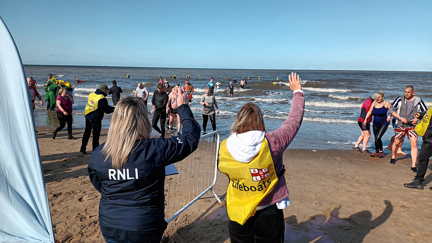 RNLI volunteers on the beach at the Rhyl Winter Dip event. Swimmers are coming out of the water. It’s a bright, sunny day with clear blue skies.