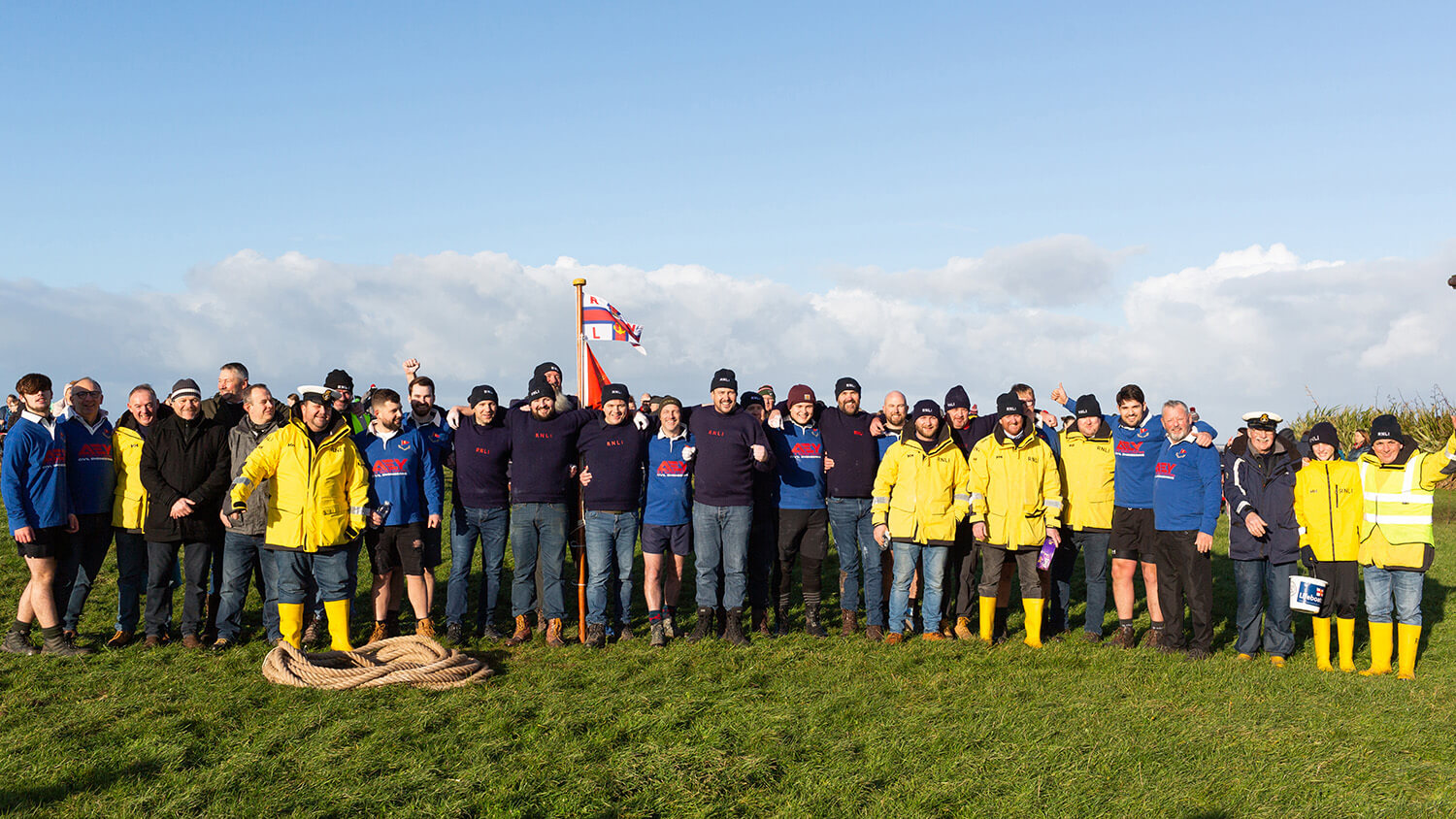 A group photo of Hoylake RNLI volunteers and members of Hoylake Sailing Club who took part in the 50th Slater Trophy Boxing Day Tug of War in 2022. They are standing outdoors on grass and the sky is blue.