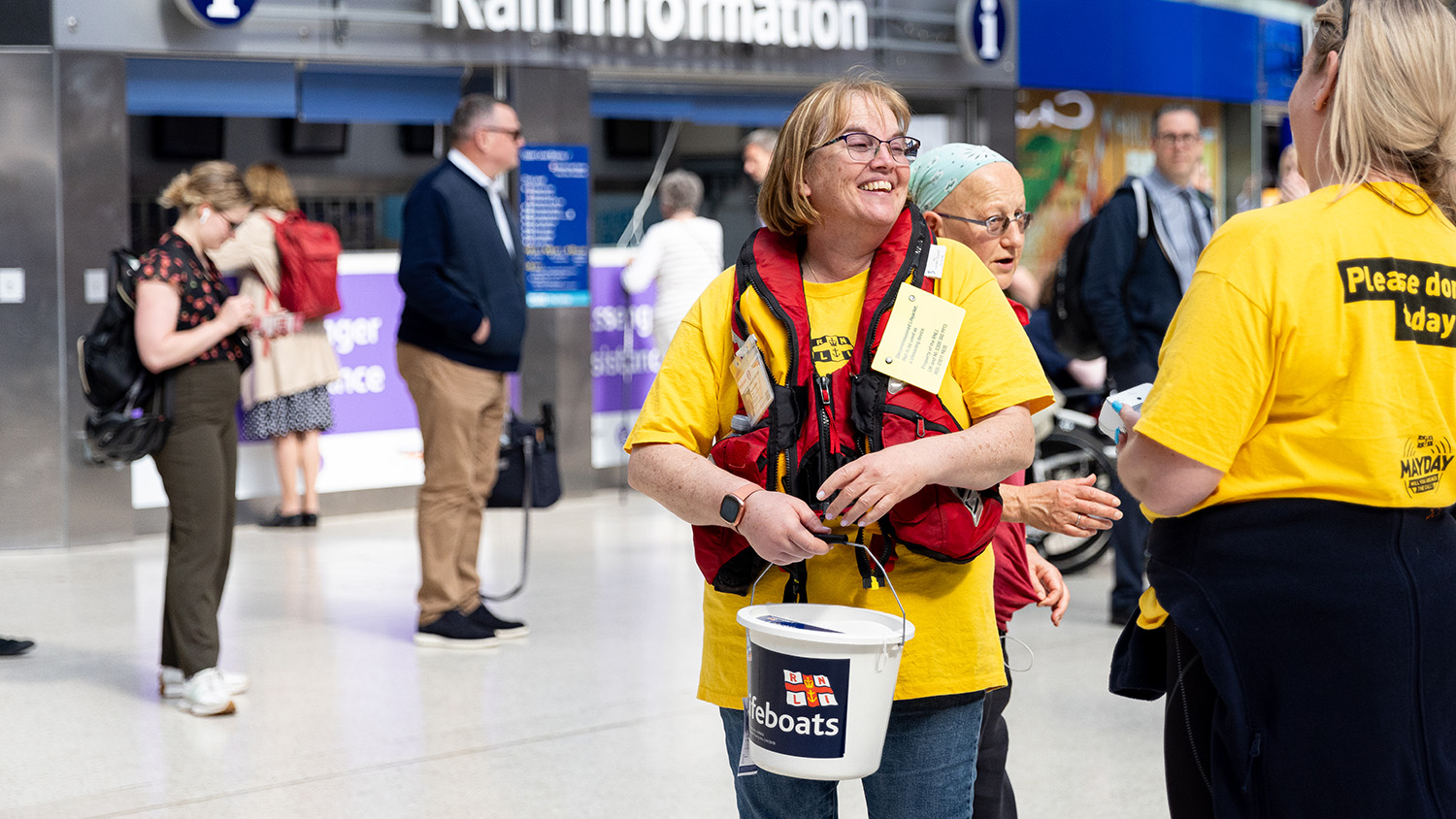 Two fundraising volunteers at a train station holding collection buckets. Members of the public are in the background.
