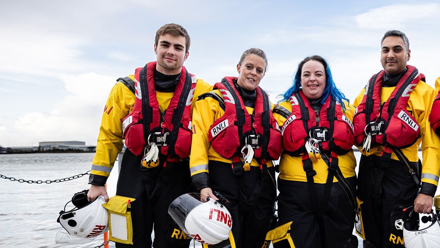 Four volunteer crew members stood in a line wearing their crew kit. Three of them are holding their helmets. The sea is in the background.
