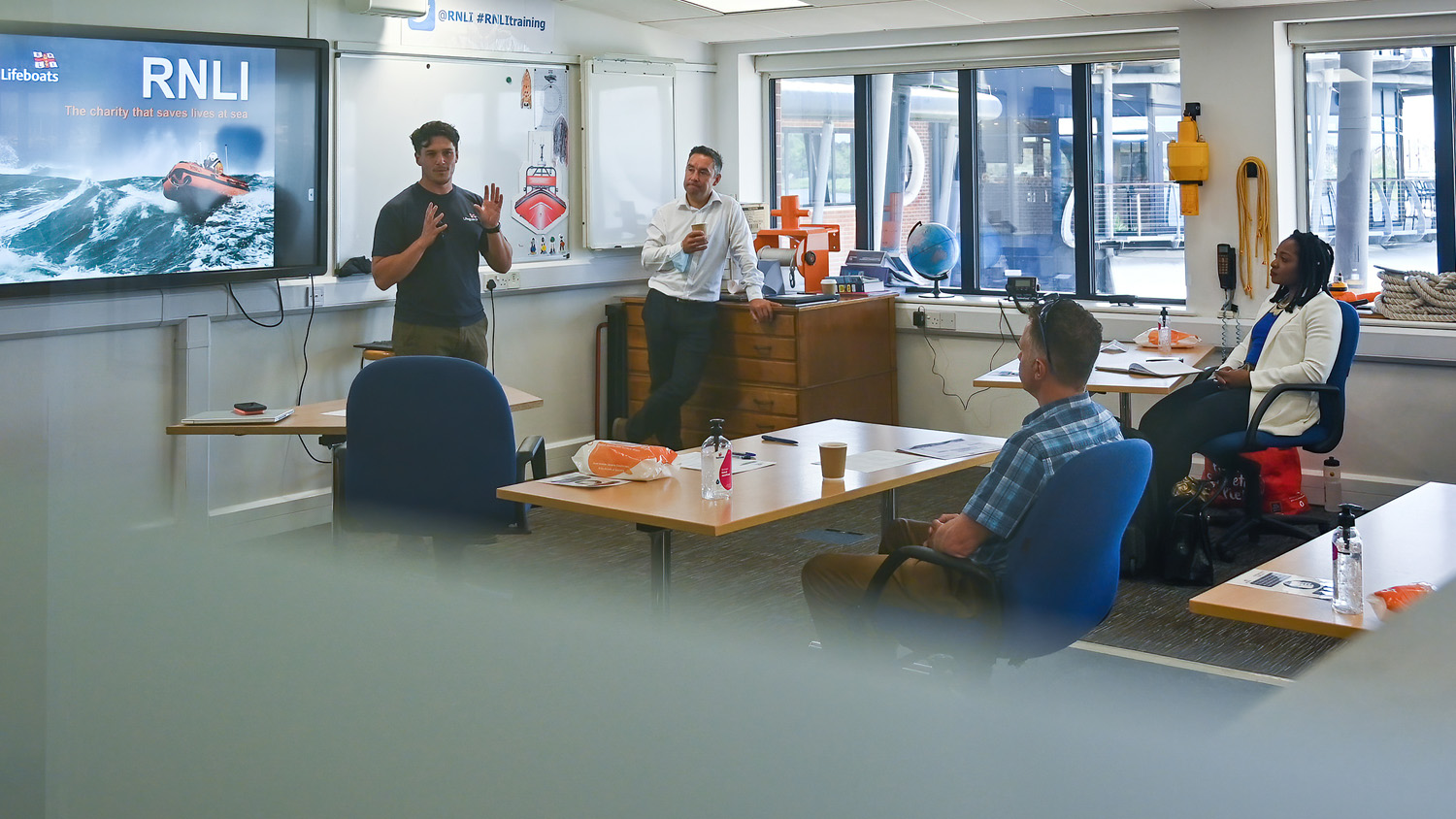 Four people are in a training room at the RNLI College. Two people are sitting at office desks, and another man stands at the side of the room. They are watching a man present some training, and there is an RNLI branded presentation on the screen behind him.