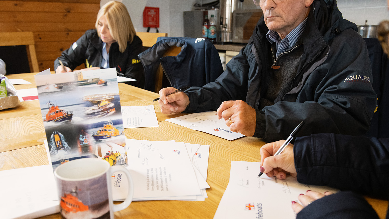 Volunteers gathered round a kitchen table writing things down on paper. On the table, there is a card with lifeboats on, as well as a mug with a lifeboat on.