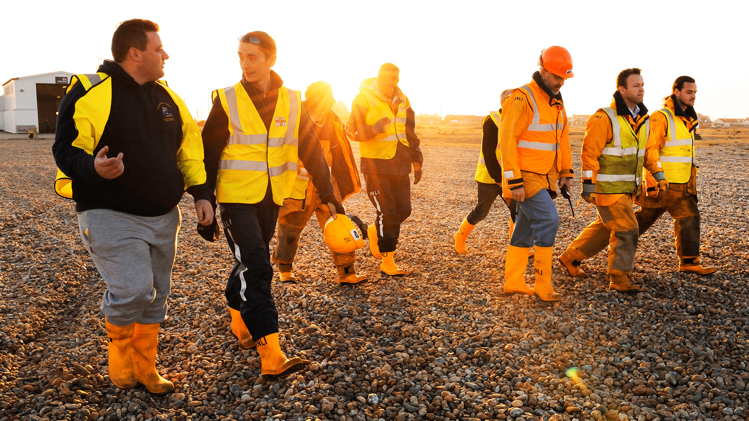 A group of lifeboat station volunteers walking along a pebble beach at sunset.