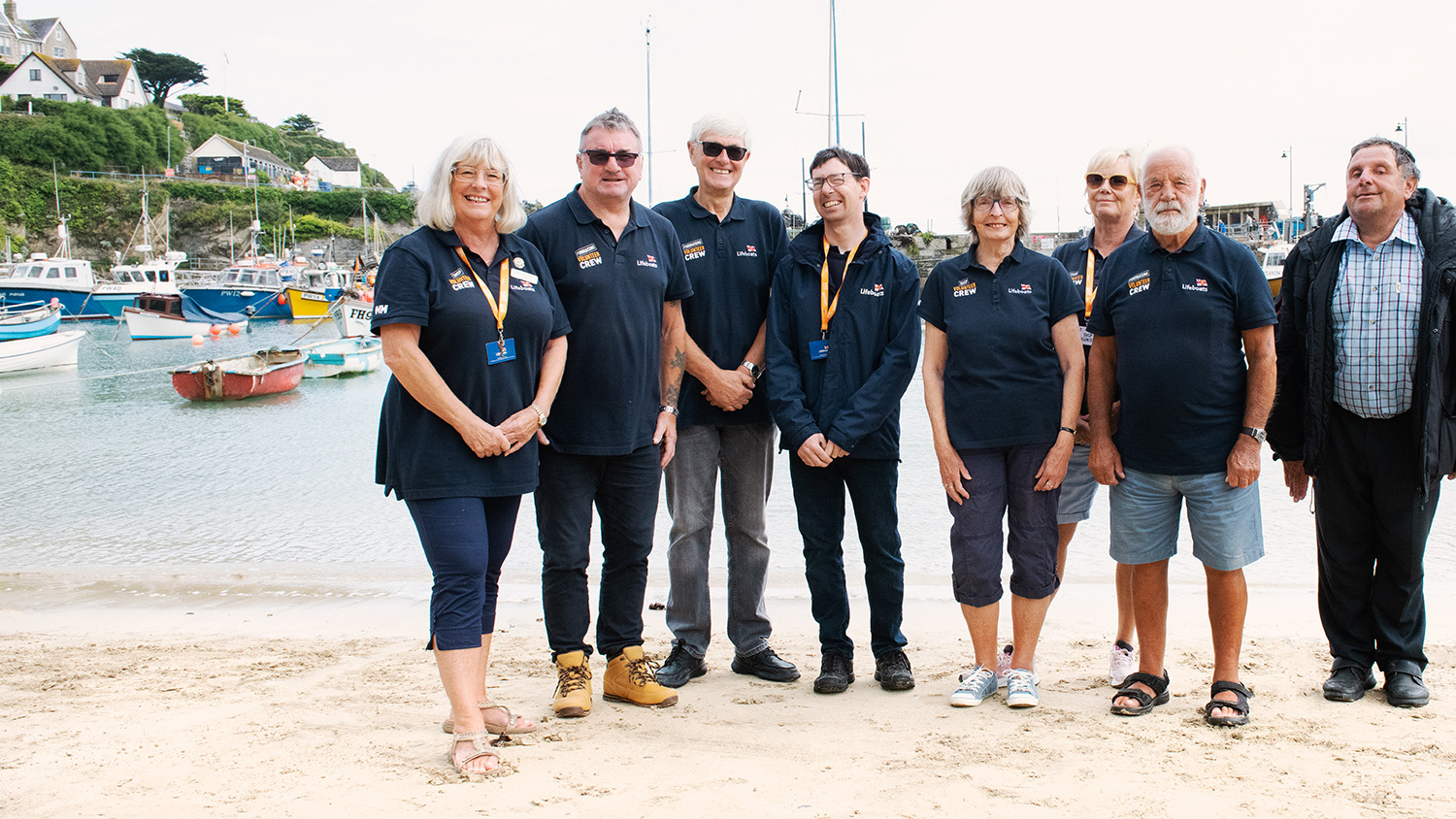 A group of volunteers wearing RNLI branded clothing stood on a sandy beach. There are boats on the water in the background.