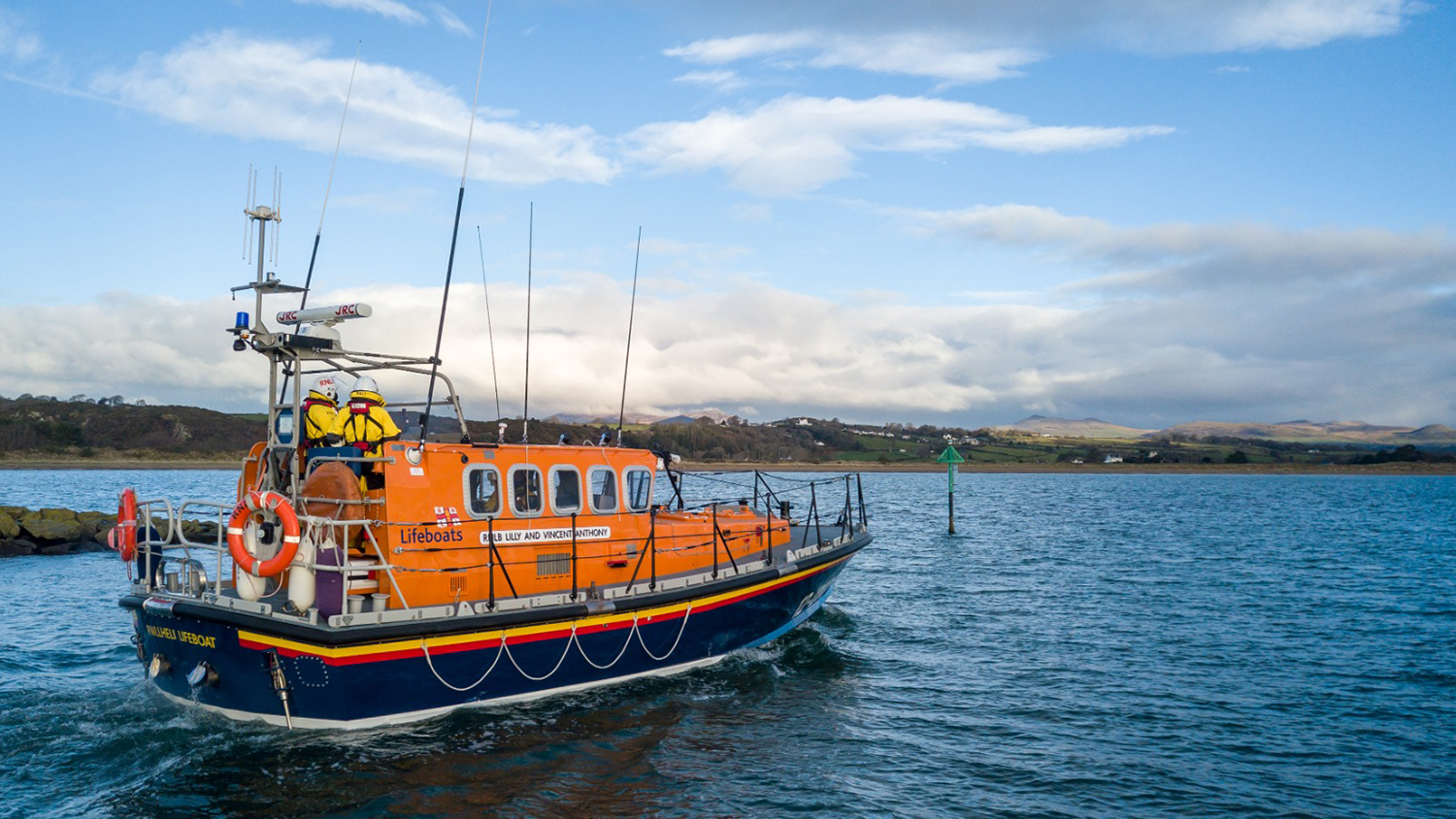 A Mersey class all-weather lifeboat is heading out to sea on a calm, sunny day. Two crew members are standing in the centre of the deck, facing the horizon. 