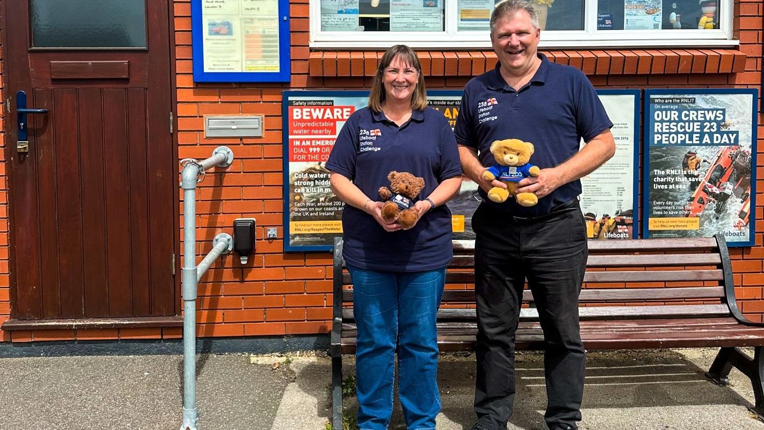 A male and female couple stand outside a lifeboat station next to a wooden bench. They are smiling at the camera and both holding an RNLI teddy bear.