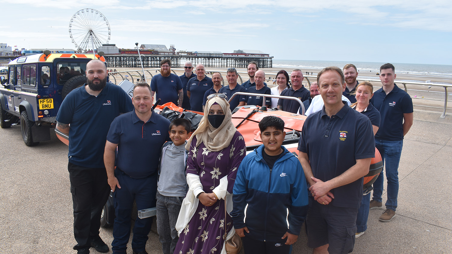 Group photo of RNLI lifeboat crew members and visitors standing beside an inshore lifeboat on Blackpool seafront, with the pier and Ferris wheel in the background.