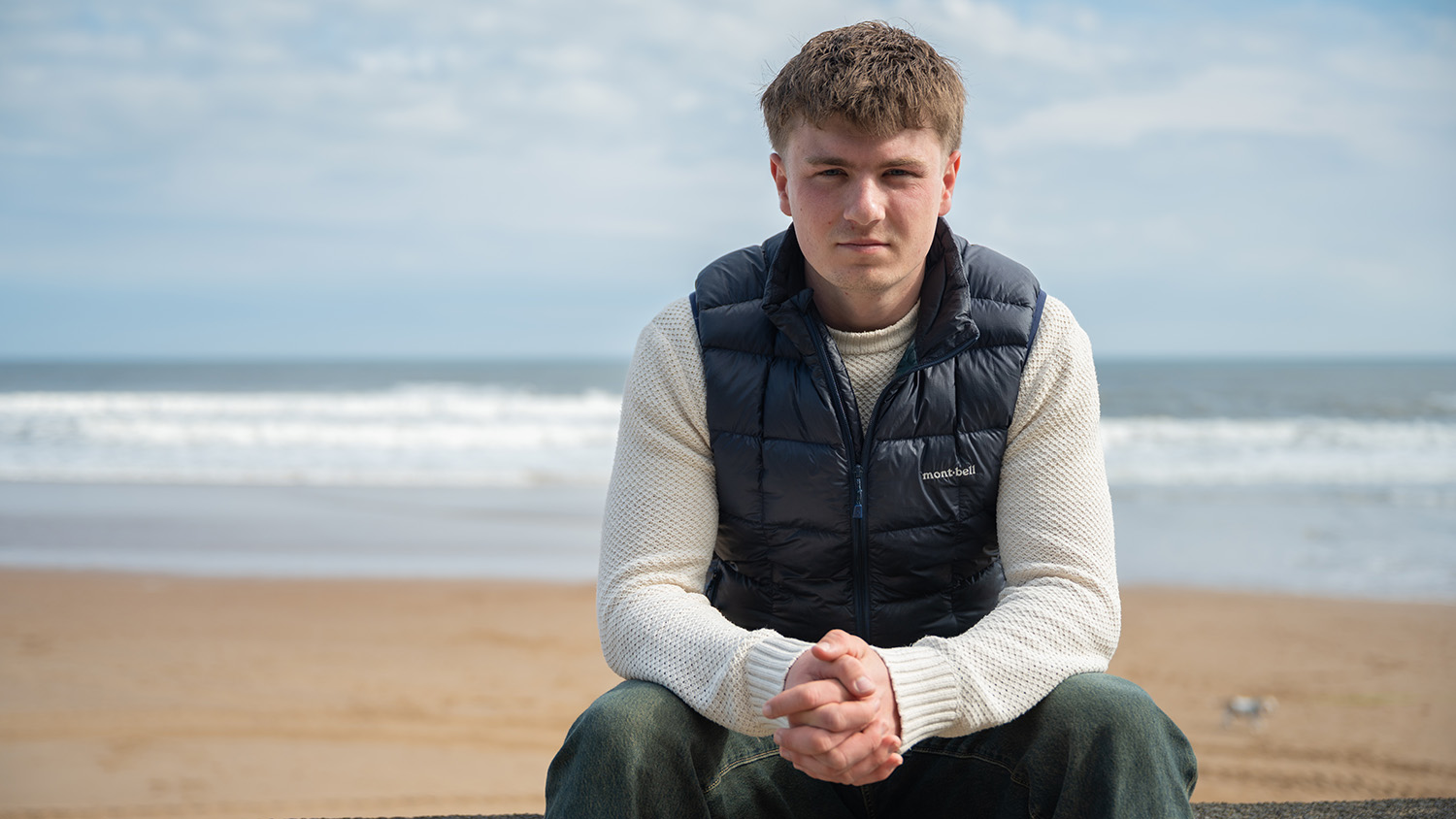 Euan sitting on a wall and leaning forwards with the beach behind him.