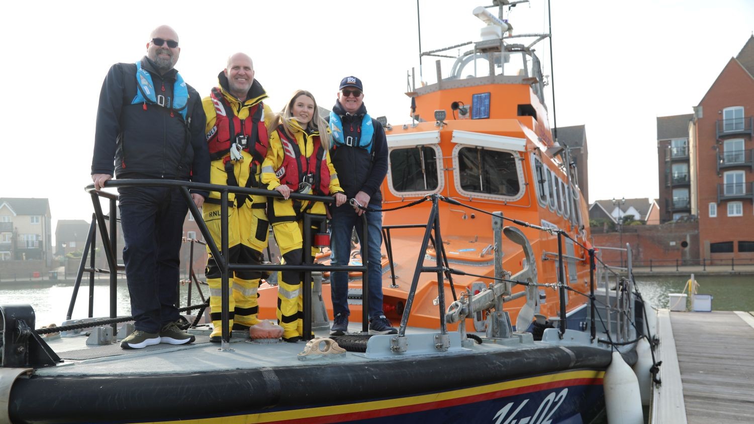 Three men and one woman standing on the front deck of an orange all-weather lifeboat