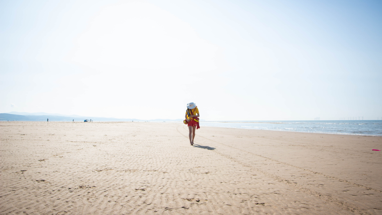 A lifeguard walking alone along a wide, empty sandy beach on a bright, clear day. The sea is visible to the right, with gentle waves along the shoreline, and the sky is pale and cloudless, creating a calm, open atmosphere.