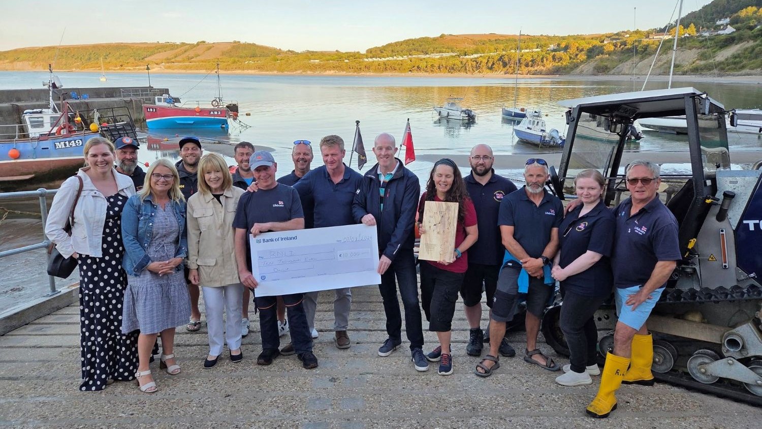 A group of people stand at the top of a boat slipway with the water behind them. Two men in the centre are holding a large cheque.