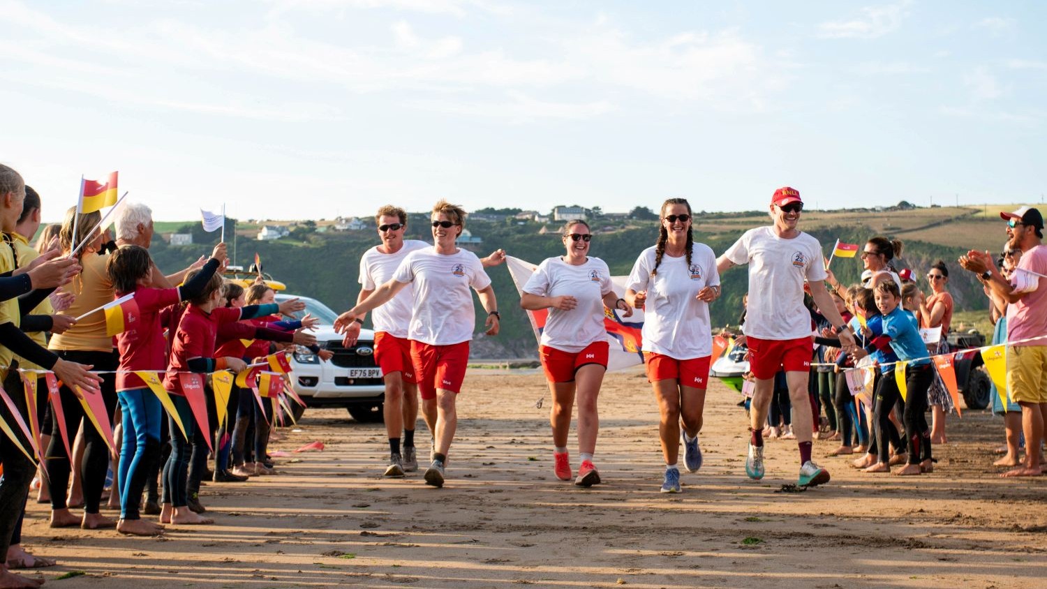 Five people in matching white tshirts and red shorts running on a beach between two rows of people cheering them on