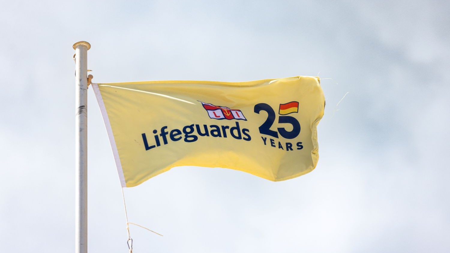 A yellow flag with Lifeguards 25 Years logo flying against a cloudy sky