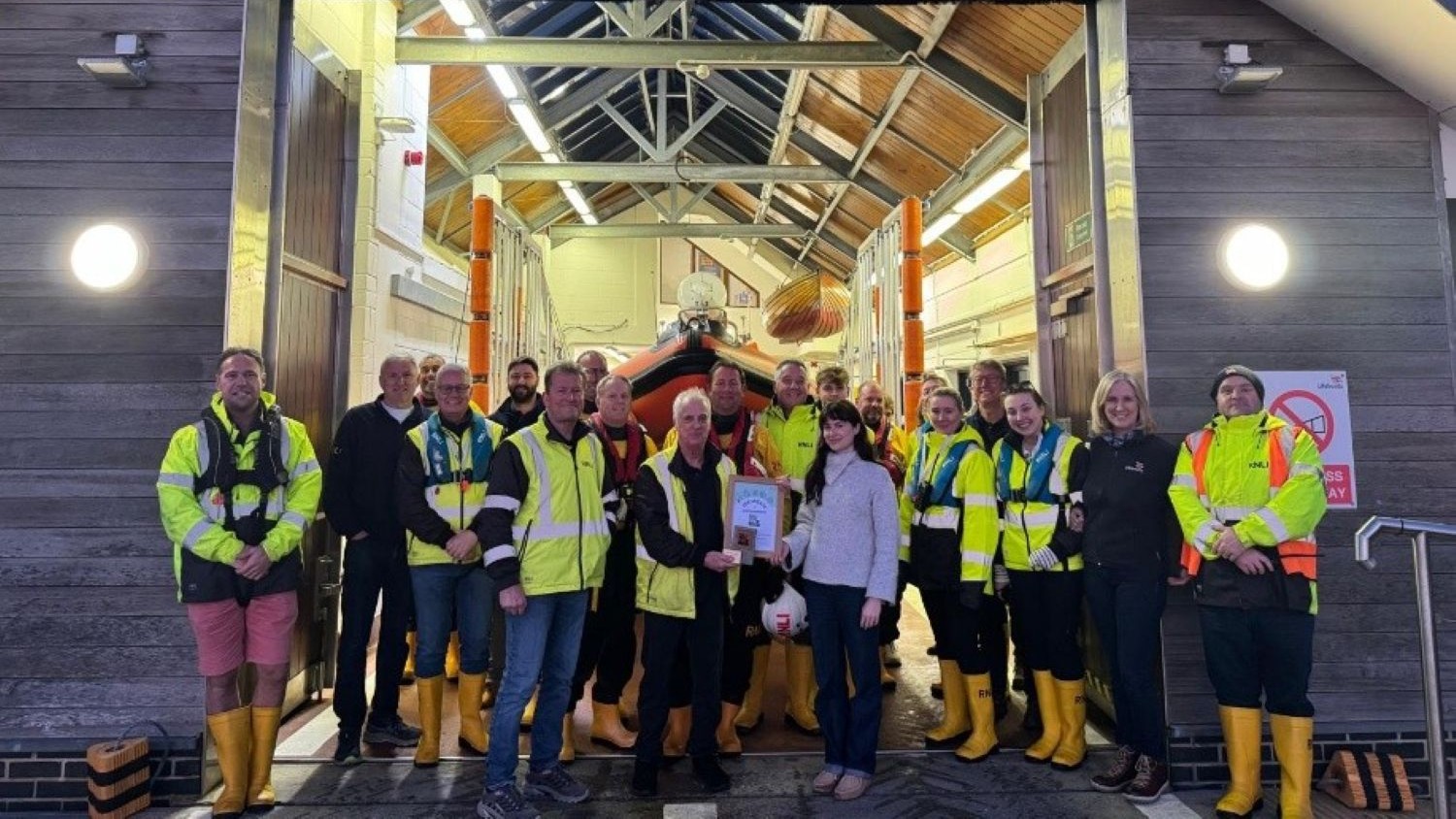 A line-up of people mostly wearing hi-vis jackets and yellow RNLI wellies. A man and a woman in the centre are shaking hands and holding a framed certificate.