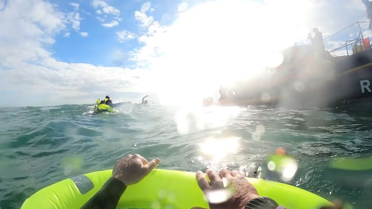 A view from a person in the sea holding onto an inflatable device while looking towards an RNLI lifeboat, with multiple other people in the water