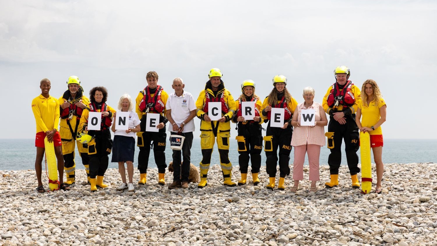 A line of volunteers including boat crew, lifeguards, retail and fundraisers standing on a pebble beach