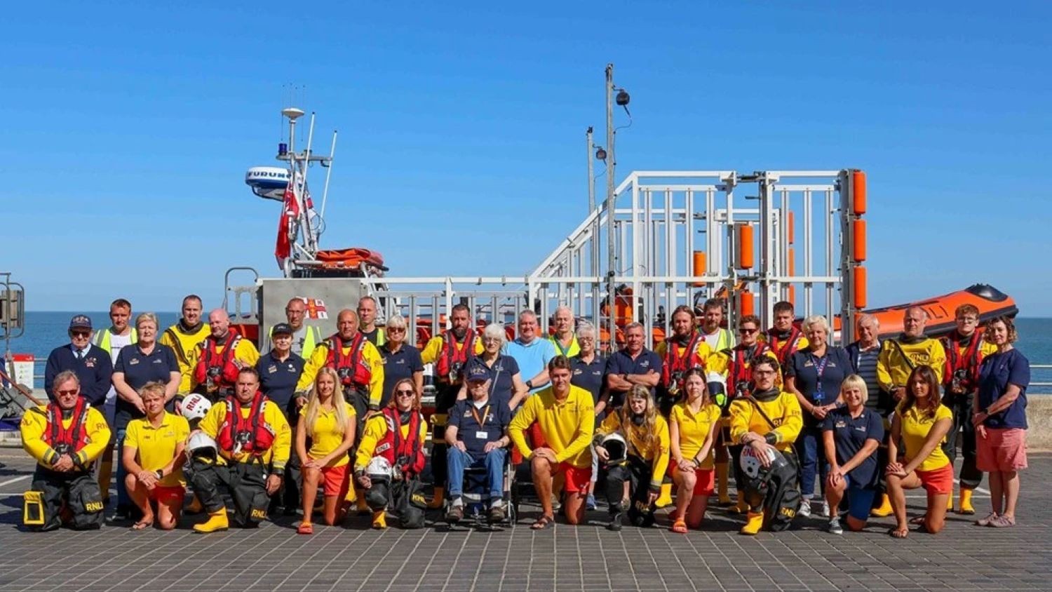 A large group of volunteers and lifeguards lined up in front of an inshore lifeboat. Blue sky and sea can be seen in the background.