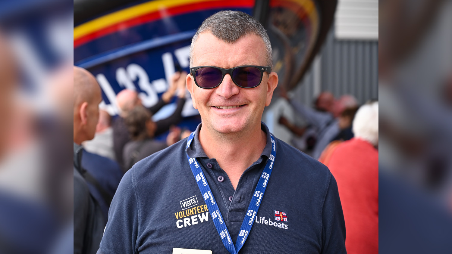RNLI volunteer Stuart Lindsay smiling and wearing a 'Visits Volunteer Crew' polo shirt, standing in front of a lifeboat.