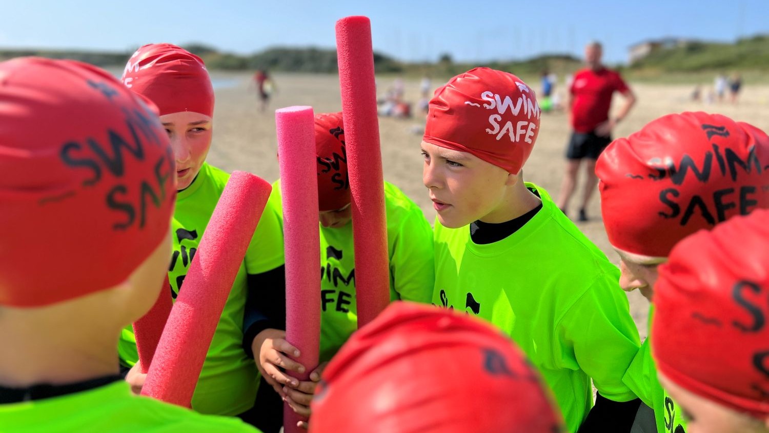 A group of young people on a beach wearing neon green tshirts and red Swim Safe branded swimming caps