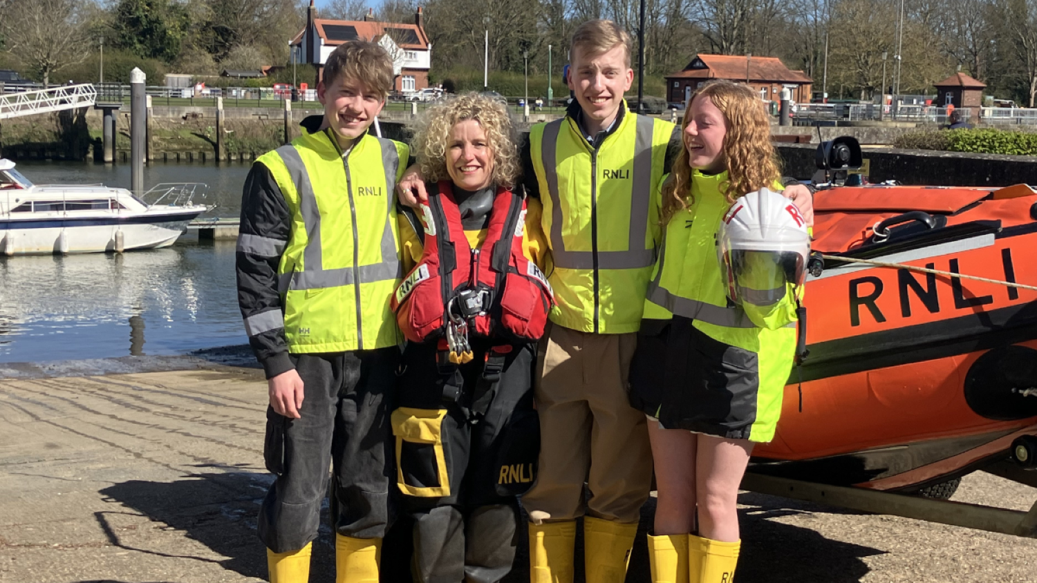 Four people lined up in front of an orange inshore lifeboat. Three are wearing yellow hi-vis vests and yellow wellies; a fourth woman wears full crew kit.